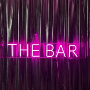 Pink neon sign saying the bar hanging against silver drape