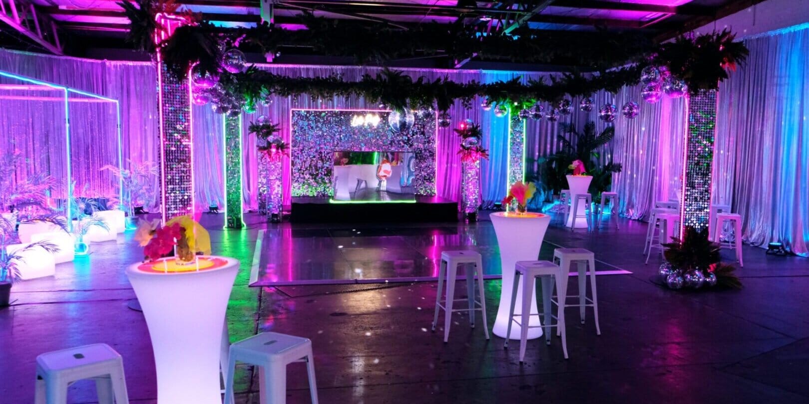 Room shot of neon disco party theme featuring illuminated furniture, bar stools, greenery, dance floor, neon lights, and sequin panels
