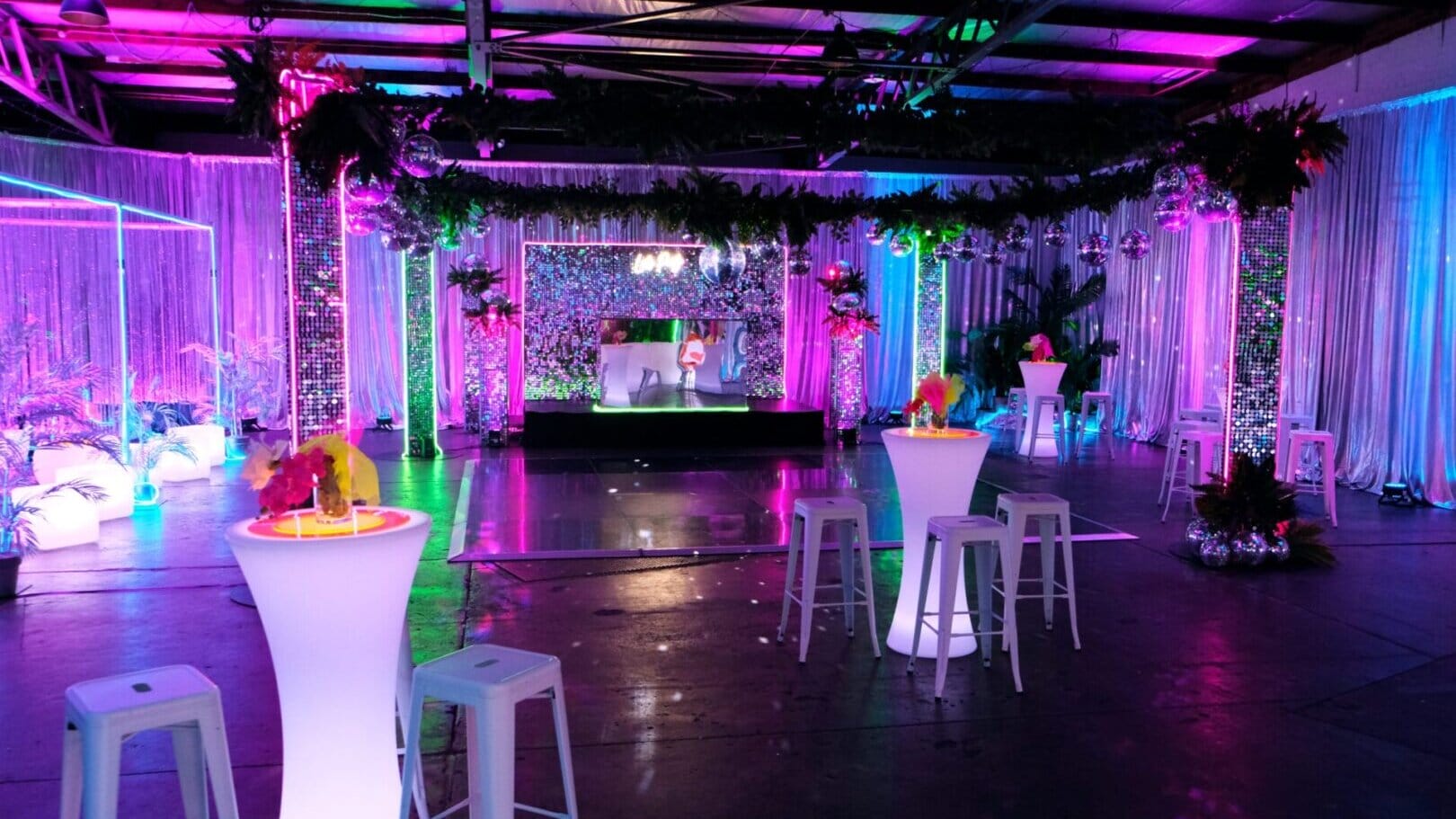 Room shot of neon disco party theme featuring illuminated furniture, bar stools, greenery, dance floor, neon lights, and sequin panels