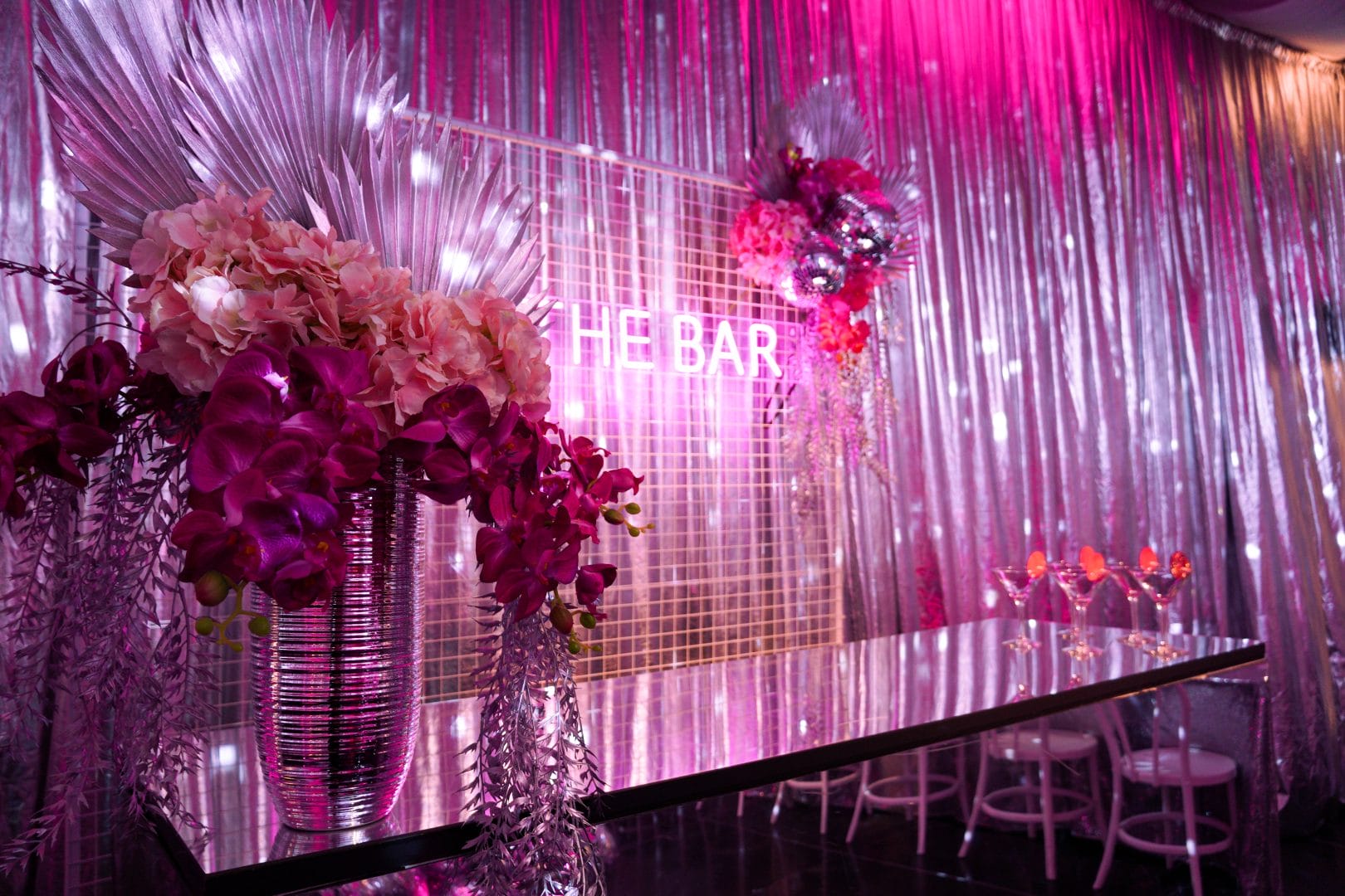 the bar at a pink disco dinner party setup. Area features a pink acrylic bar, florals, a mesh backdrop, and a pink neon 'the bar' sign