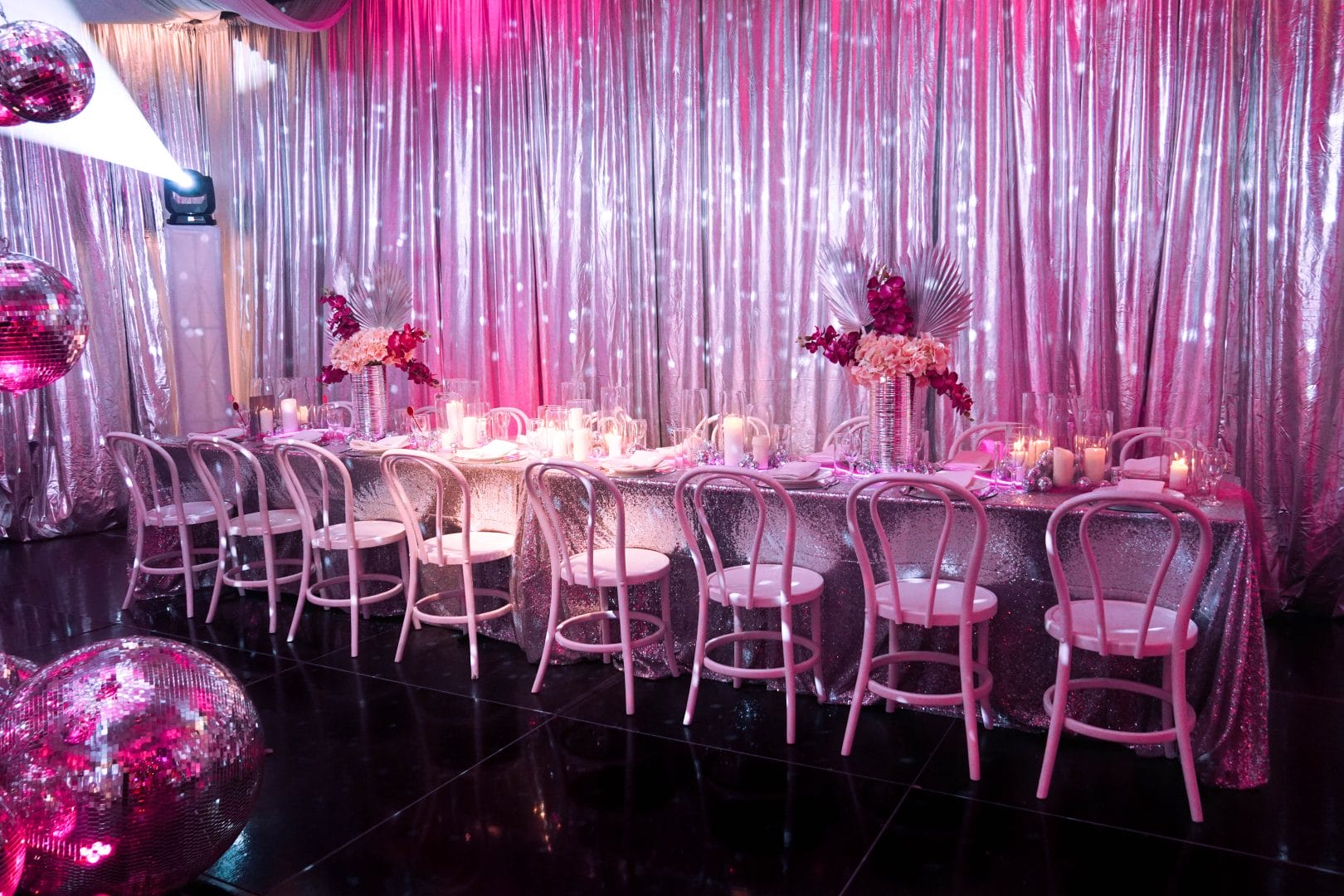 Long table setup featuring pink and silver decor, mirror balls, silver drape, pink lighting
