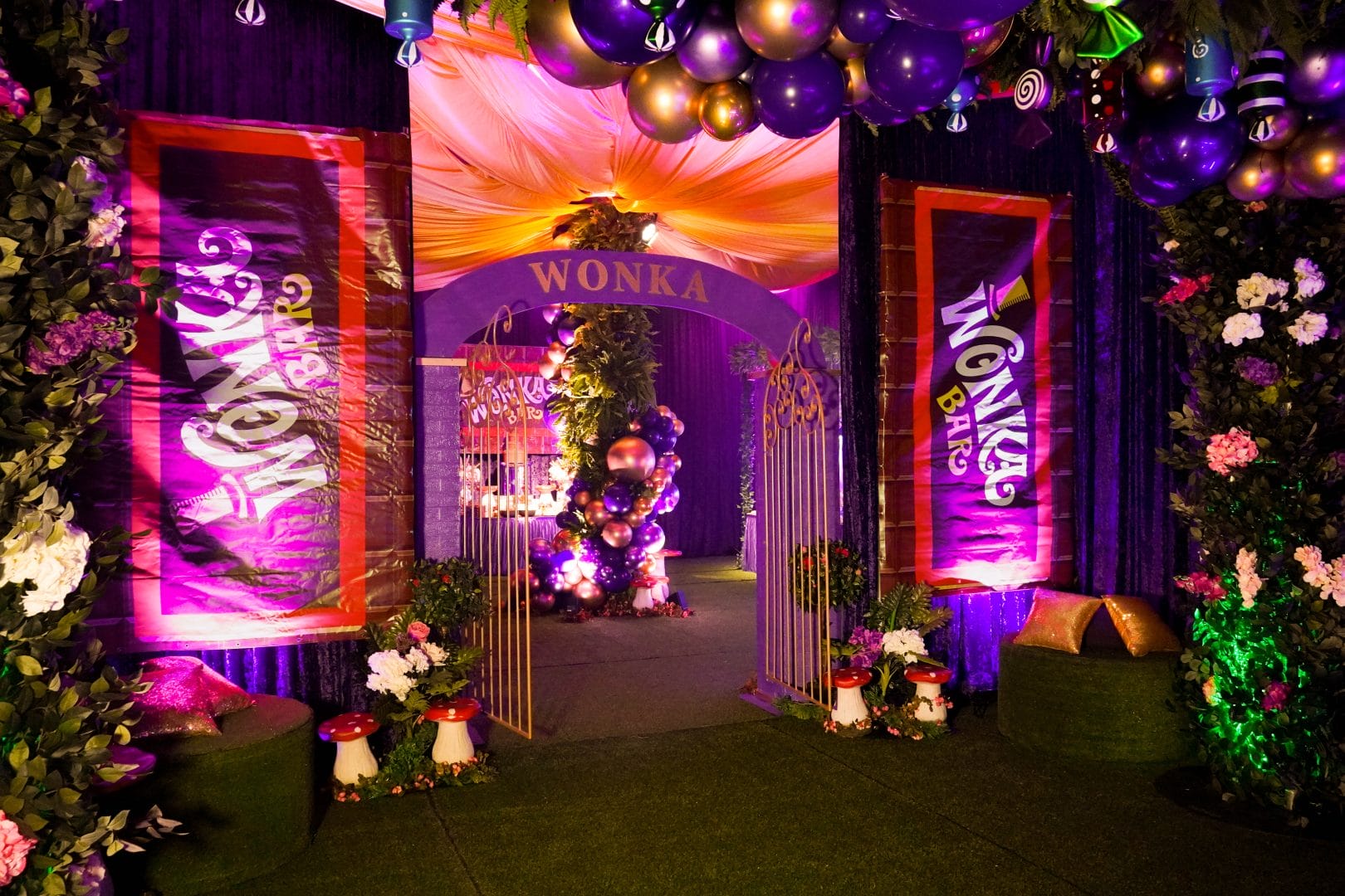 Wonka candyland themed entrance to the dance floor area featuring balloons, themed banners, lots of greenery, and flowers