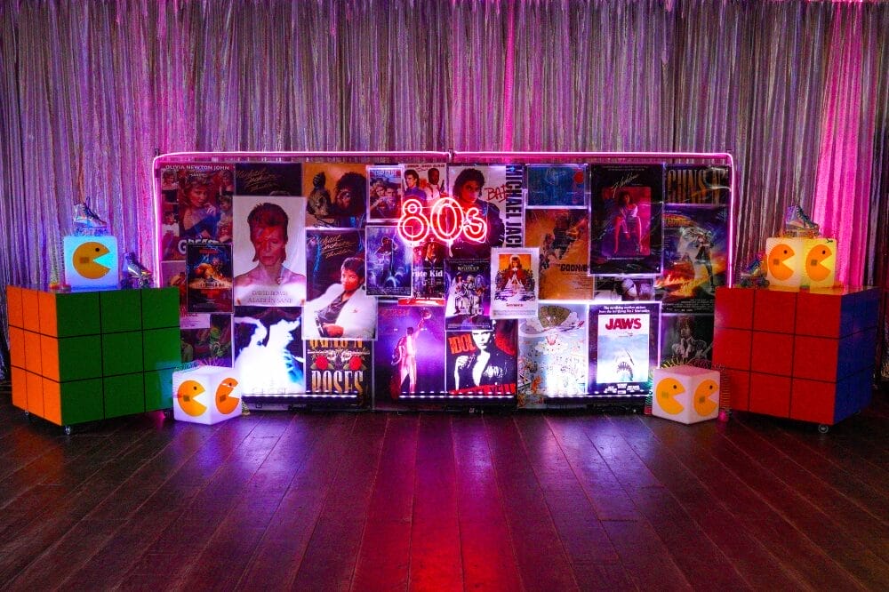 80s themed backdrop with a neon sign, 80s movie posters, and themed decorating items